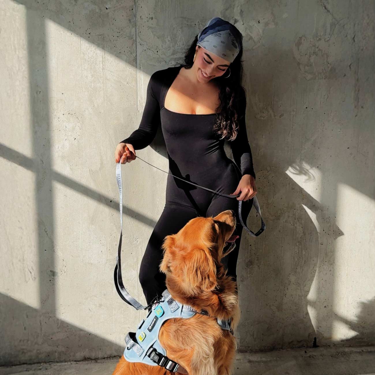 Woman in black outfit standing with a dog against a concrete wall, and the dog is wearing dusk blue Classic MagEase harness with velcro patches.
